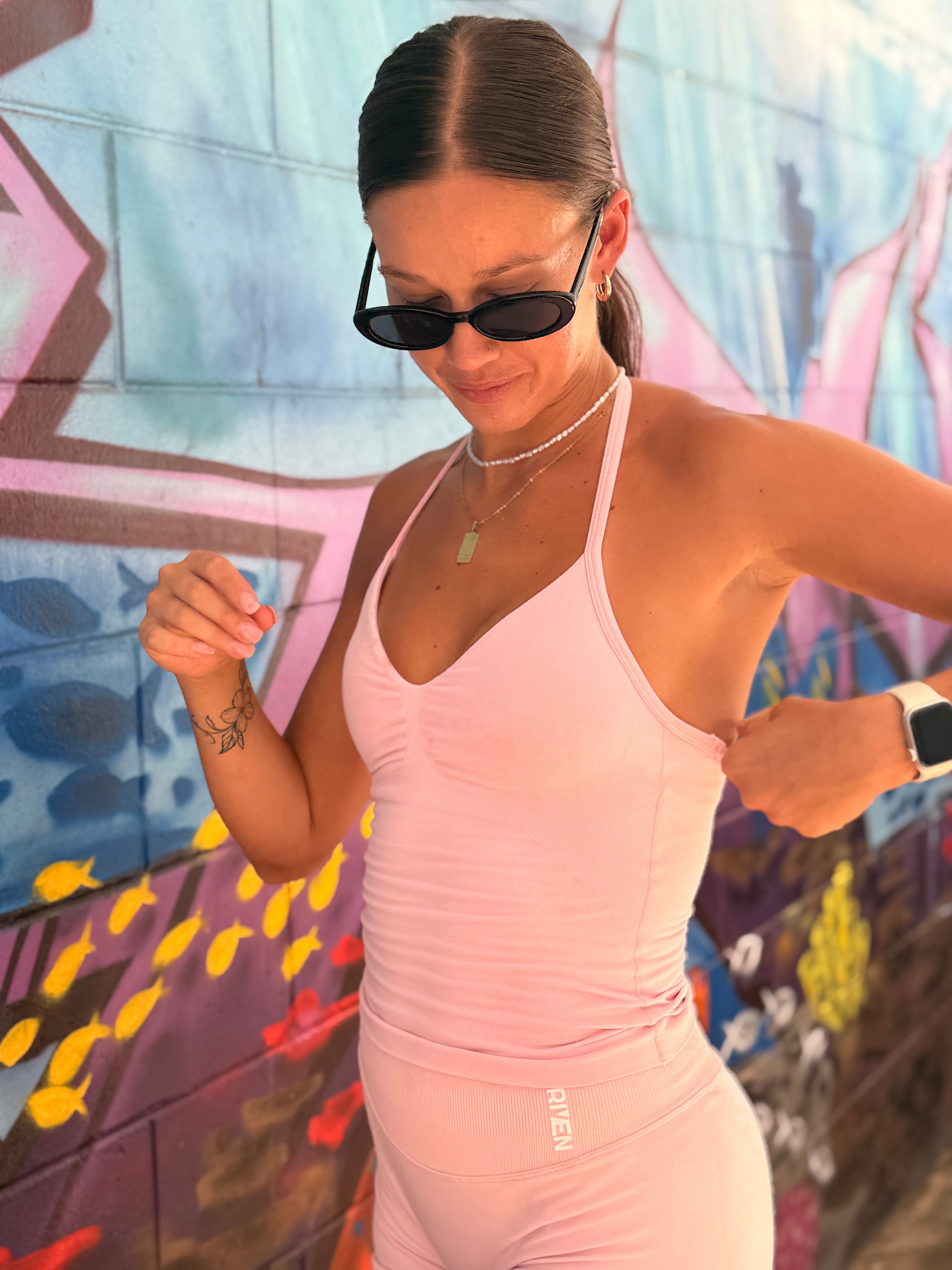 A person with long blond hair in a ponytail is wearing a brown sports bra and matching leggings. They are smiling and posing confidently with one hand resting on their hip and the other by their side. The background is plain white.